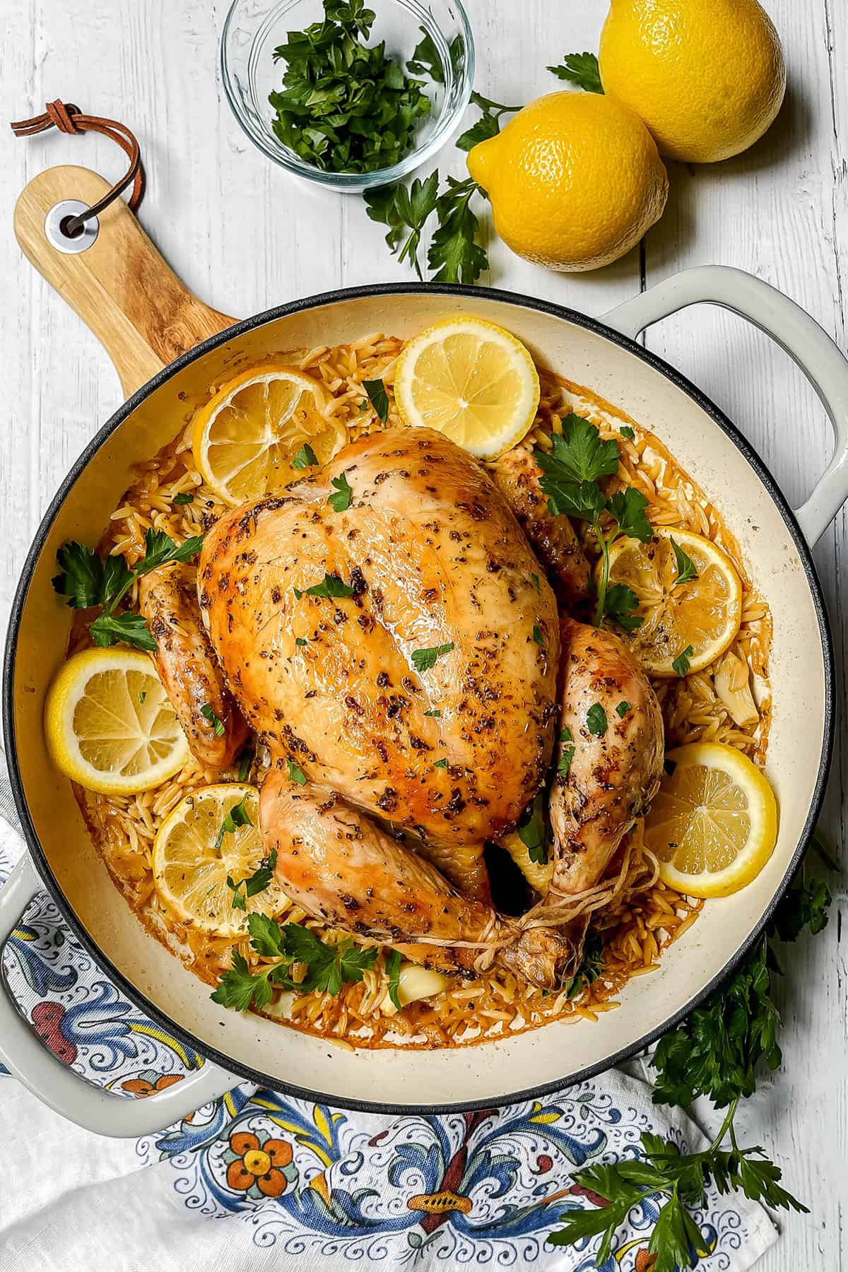 A roasted chicken sits in a pan with orzo, lemon slices, and fresh parsley; two lemons and a bowl of chopped parsley are nearby on a white wooden surface. This inviting roasted chicken dish is perfect for any meal.