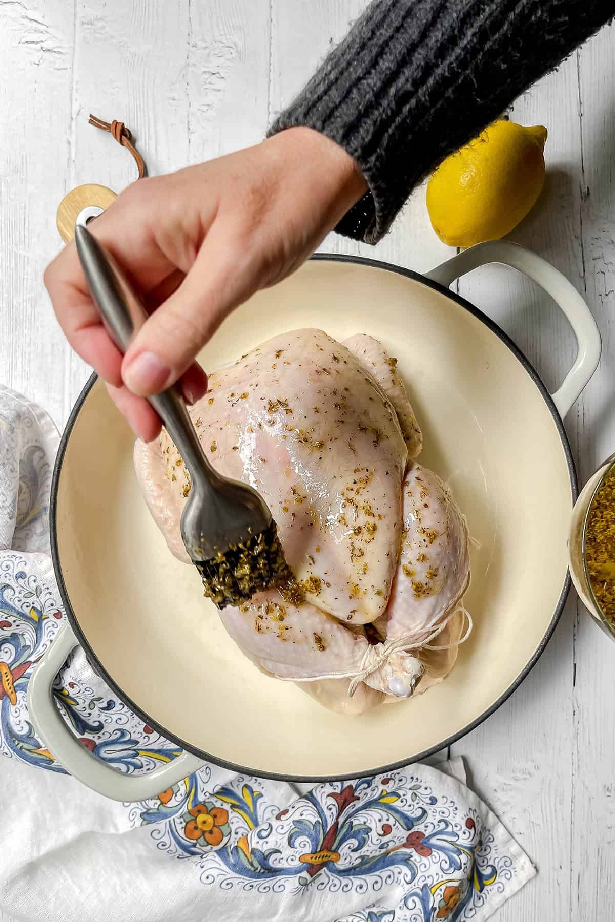 A hand brushes seasoning onto a raw whole chicken, soon to become a delicious roasted chicken, placed in a white Dutch oven. A lemon and a bowl of seasoning sit nearby on the white surface.