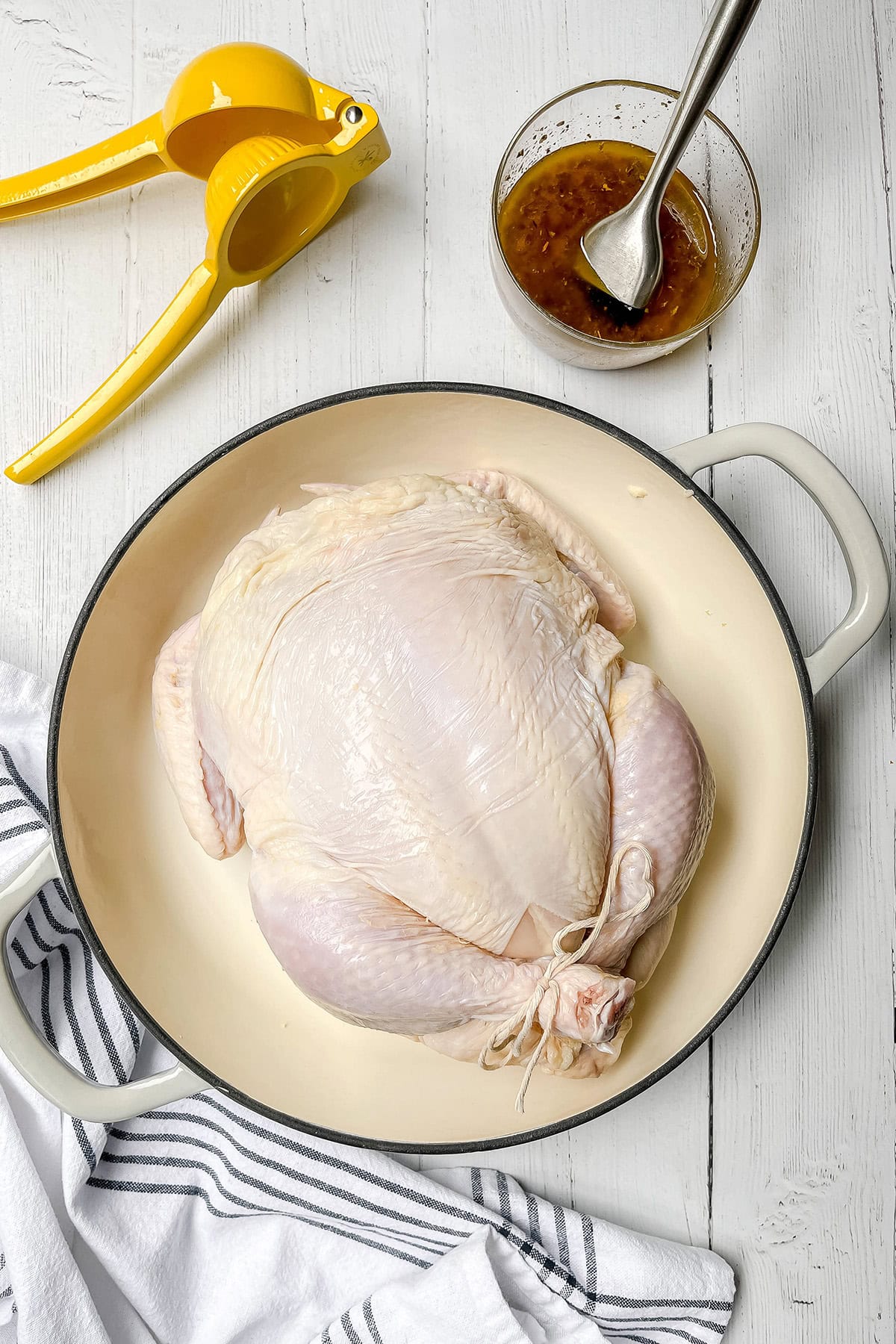 Raw whole chicken in a white baking dish, ready to become delicious roasted chicken. Surrounded by a citrus juicer, a bowl of marinade, and a striped towel on a white wooden surface.