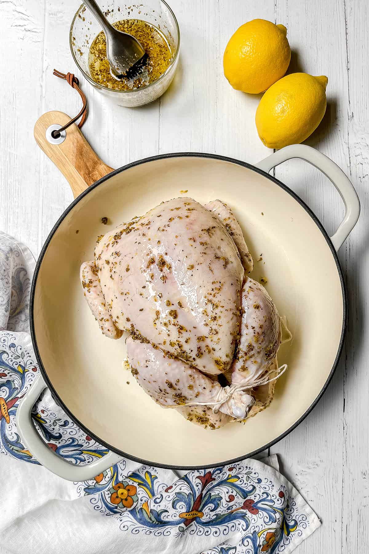 Raw whole chicken seasoned with herbs in a white roasting pan, ready to become delicious Roasted Chicken, with two lemons and a bowl of marinade or dressing on a white wooden surface.