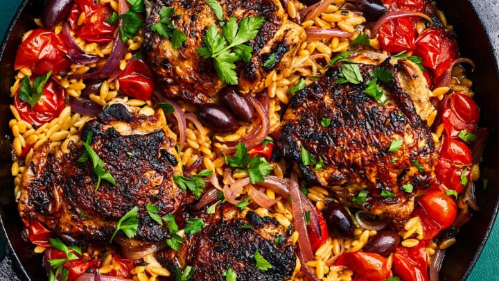 A cast iron skillet filled with seared chicken thighs, orzo pasta, cherry tomatoes, red onions, olives, and parsley—perfect for cozy January One Pot Dinners.