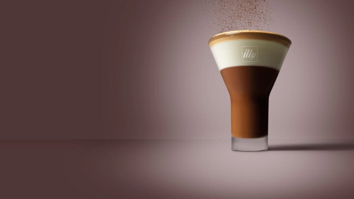 A glass filled with layered espresso and cream, topped with cocoa powder, sits on a plain brown surface with a blurred brown background—an inviting addition to your favorite winter coffee recipes.