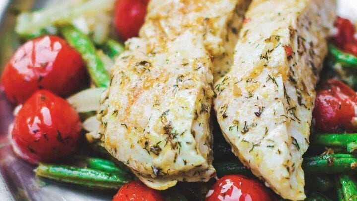 Two fillets of seasoned, cooked white fish served on a metal plate with green beans, cherry tomatoes, and onions—perfect for those following the Mediterranean Diet.