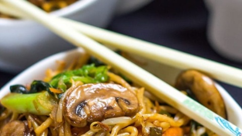 A bowl of Chinese New Year noodles stir-fried with mushrooms, carrots, and greens, served with chopsticks. Another bowl is visible in the background.
