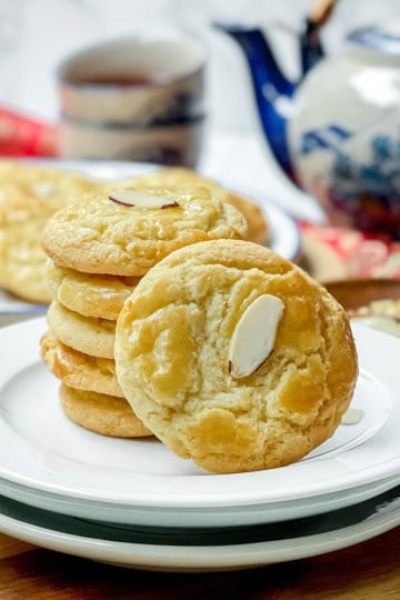 A stack of Chinese Almond Cookies and a single cookie on white plates, with a teapot and cup in the background on a wooden table.