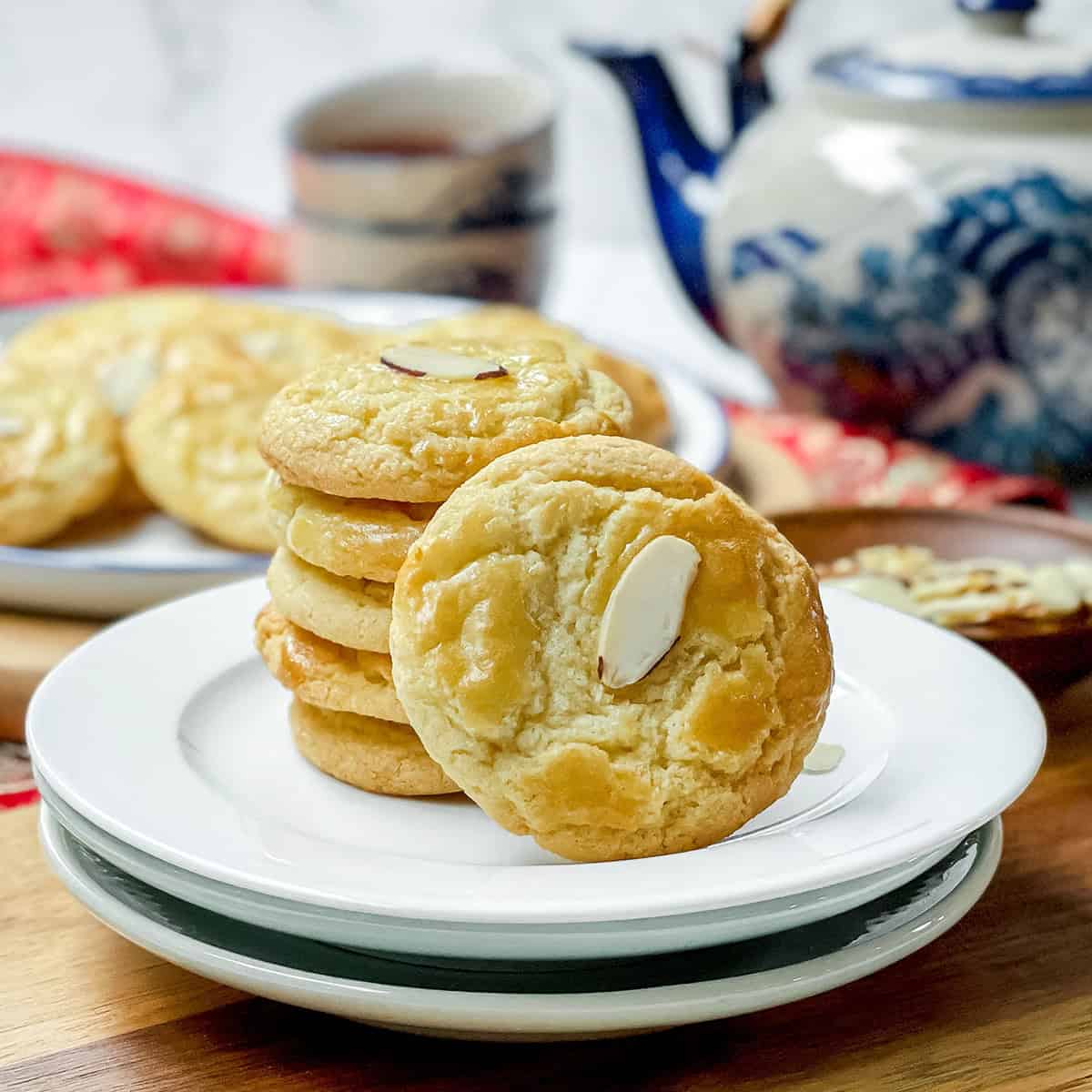 A stack of Chinese Almond Cookies and a single cookie on white plates, with a teapot and cup in the background on a wooden table.