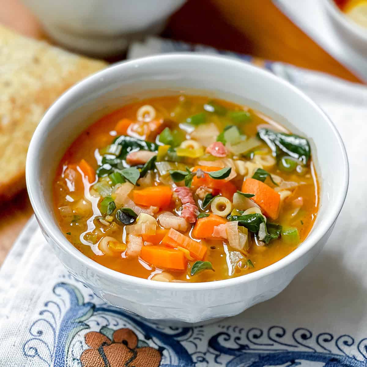 Square image of Italian Minestrone Soup with Crispy Pancetta in a white bowl with vegetables and greens.