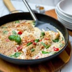 A skillet with creamy Tuscan Chicken breasts, crispy bacon pieces, red pepper slices, and fresh basil leaves, with a spatula resting inside the pan.