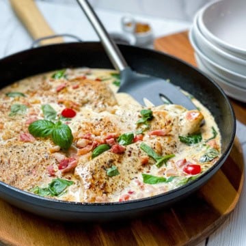 A skillet with creamy Tuscan Chicken breasts, crispy bacon pieces, red pepper slices, and fresh basil leaves, with a spatula resting inside the pan.