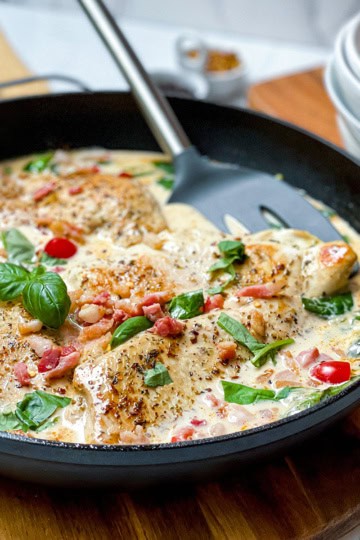 A skillet with creamy Tuscan Chicken breasts, crispy bacon pieces, red pepper slices, and fresh basil leaves, with a spatula resting inside the pan.