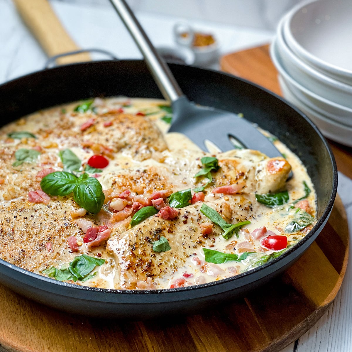 A skillet with creamy Tuscan Chicken breasts, crispy bacon pieces, red pepper slices, and fresh basil leaves, with a spatula resting inside the pan.