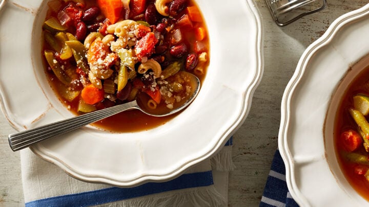 Two bowls of vegetable soup with pasta, beans, carrots, and green beans, topped with grated cheese—perfect for cozy January One Pot Dinners. A salt shaker, a wedge of cheese, and a small bowl of salt are nearby.