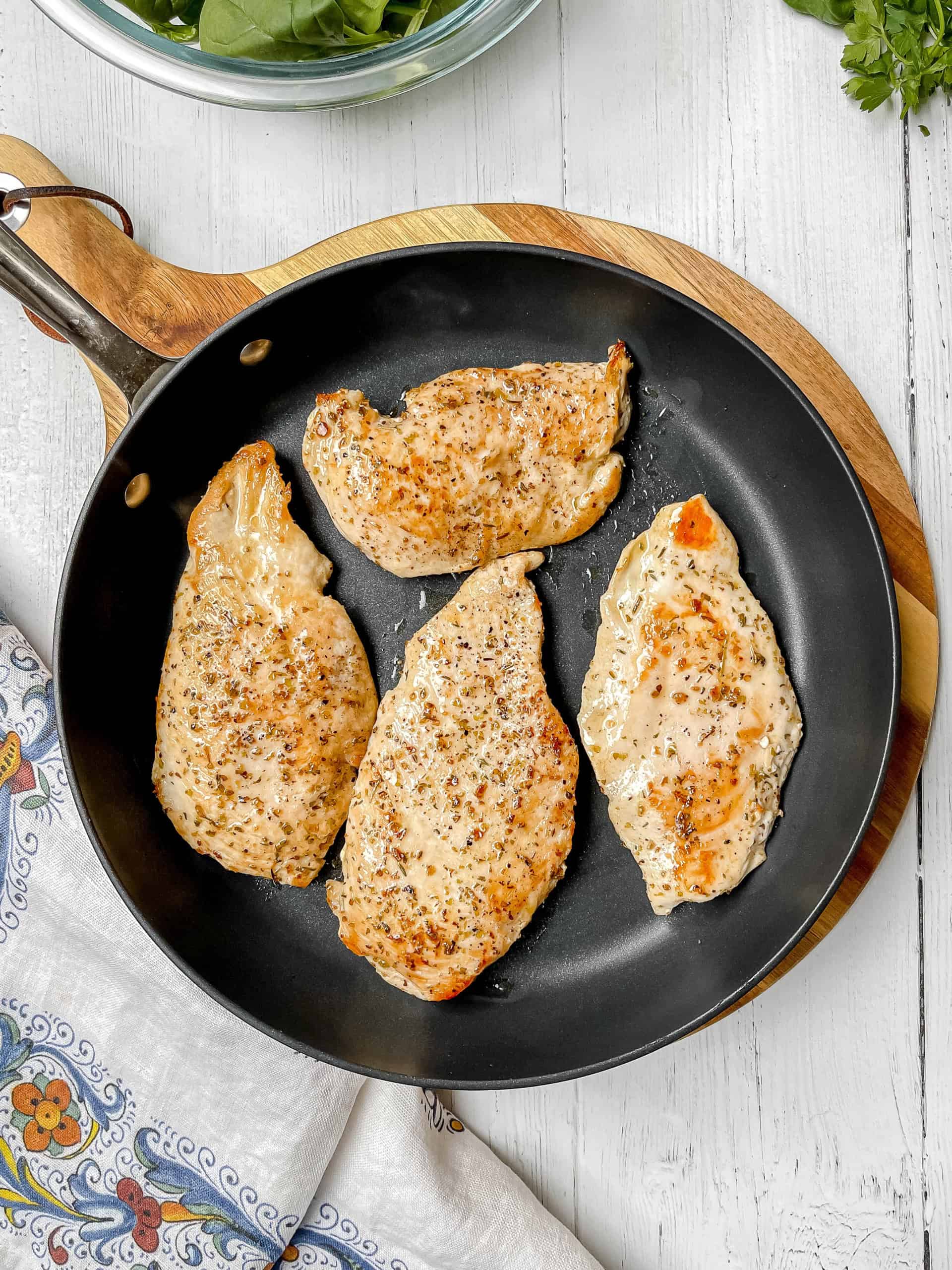 Four seasoned Tuscan Chicken breasts are cooking in a black skillet, placed on a wooden board with a cloth napkin and a bowl of greens nearby.