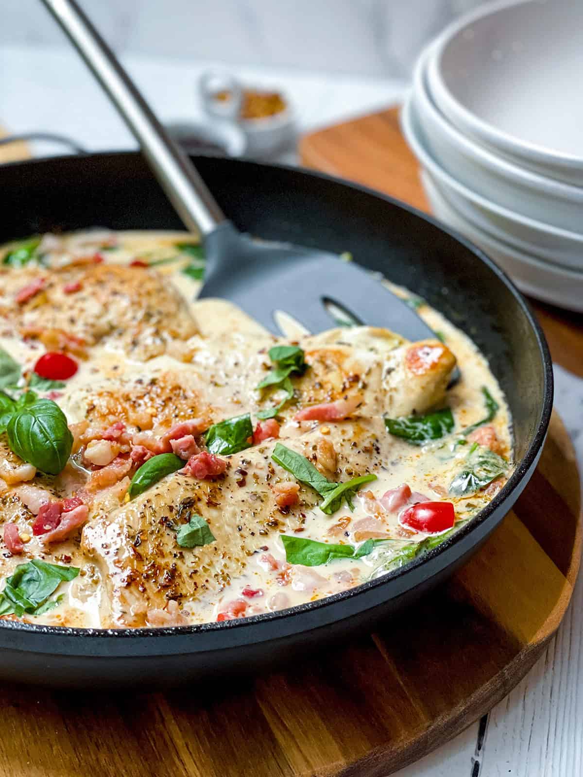 A skillet with creamy Tuscan Chicken breasts, topped with bacon, cherry tomatoes, and fresh basil, sits on a wooden board next to a stack of white plates.