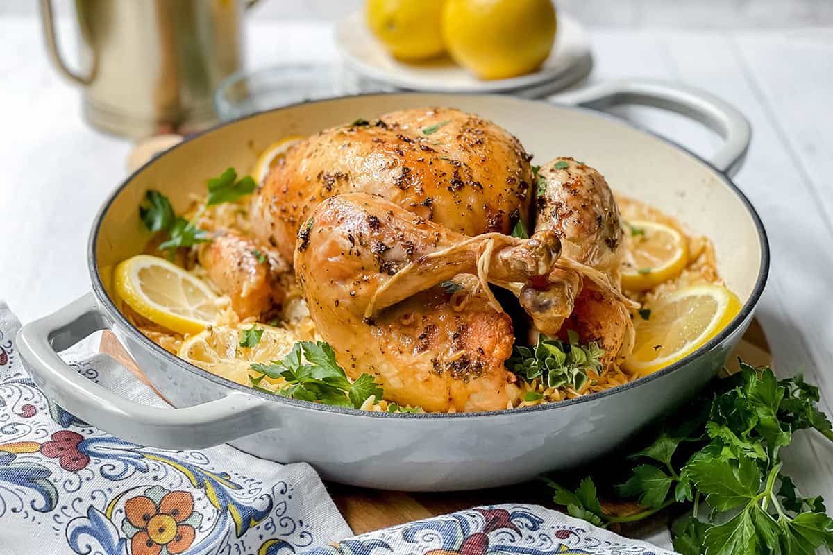 Greek roasted chicken with lemon orzo in a braising pan with lemon slices and parsley.