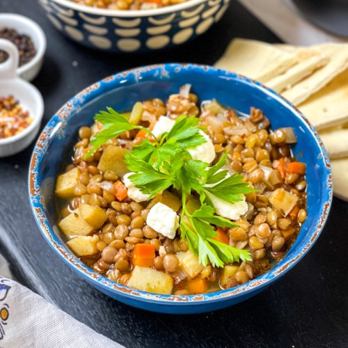 Side view of Greek Lentil Soup topped with feta and fresh parsley in a blue bowl with pita