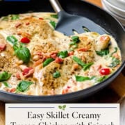 A skillet filled with creamy Tuscan Chicken, spinach, cherry tomatoes, and herbs in a rich sauce sits beside a stack of plates.