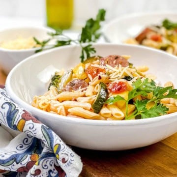 Square image of easy sausage pasta in a white bowl with vegetables and parsley garnish.
