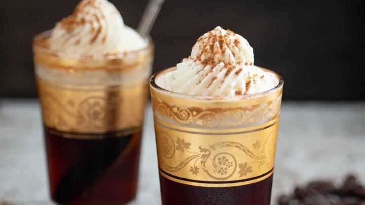 Two glasses of dark coffee topped with whipped cream and sprinkled spices, garnished with gold designs, evoke cozy winter coffee recipes. Coffee beans and spices are scattered on a wooden surface for a warm seasonal touch.