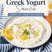 A plate of Dill Yogurt Sauce garnished with lemon slices, olive oil, and fresh herbs sits on a striped napkin, with a lemon wedge and spoon nearby.