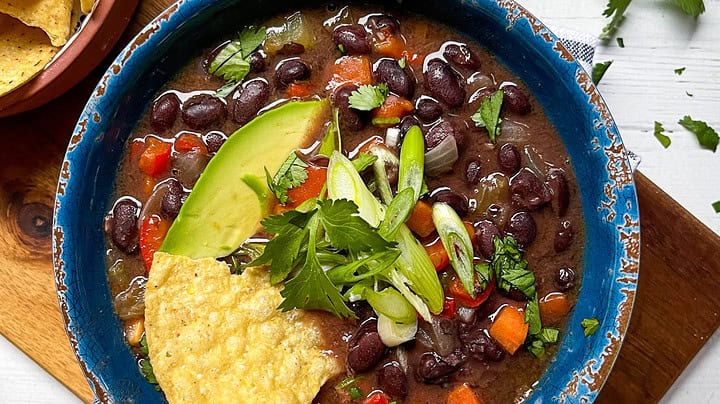 A bowl of black bean soup garnished with avocado slices, green onions, cilantro, and tortilla chips—perfect for February One Pot Dinners—sits beside a halved avocado and extra chips on a white wooden surface.