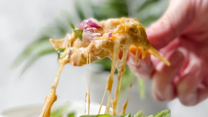 A hand lifts a cheesy portion of Black Eyed Peas Dip, topped with chopped green onions and red onions, from a dish.