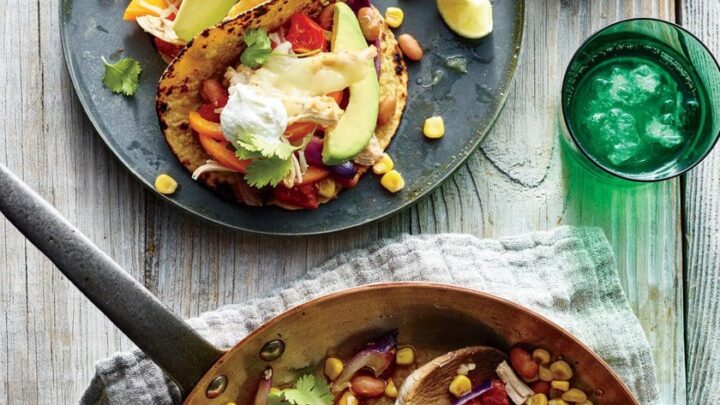 Enjoy Healthy 20 Minute Dinners with two tacos filled with vegetables, beans, avocado, and sour cream, served beside a skillet of chicken and veggies, lime wedges, and a green glass of water on a wooden table.