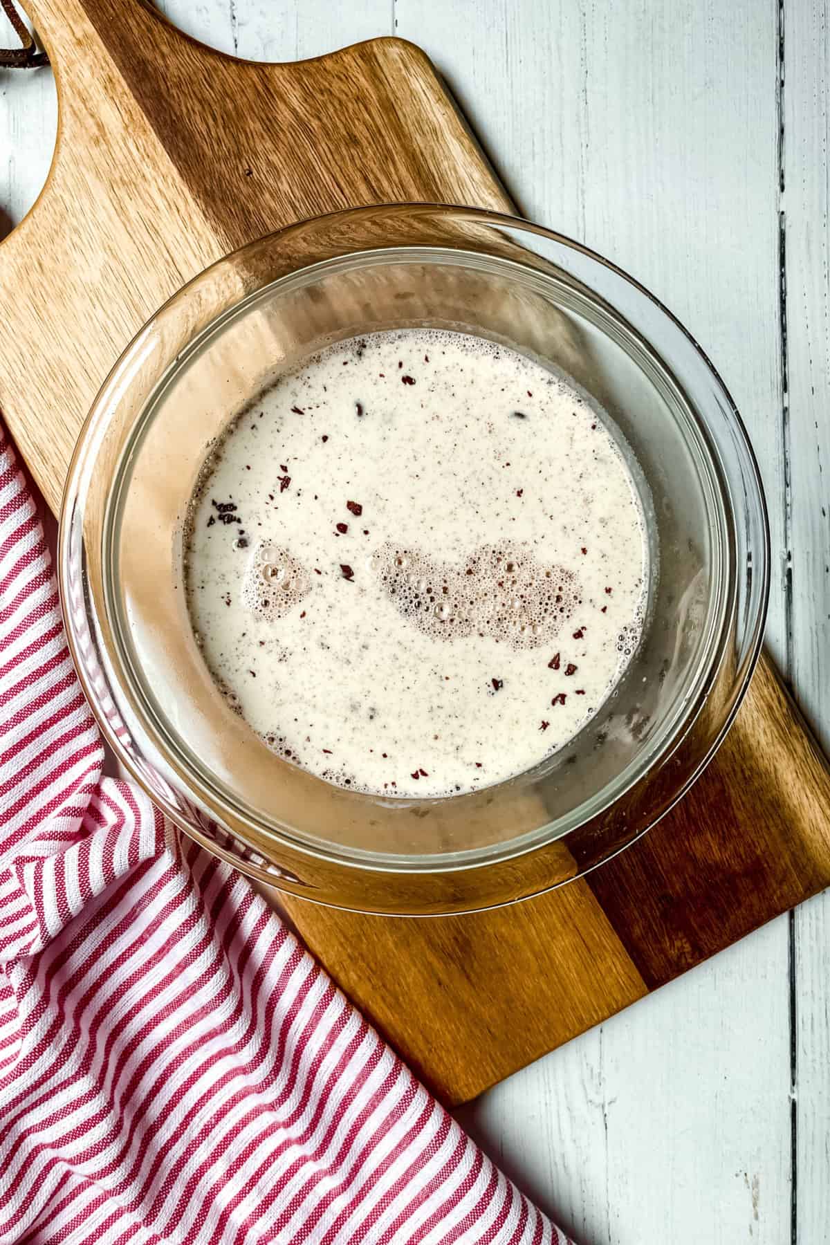 A glass bowl filled with chocolate cake wet ingredients, perfect for a chocolate cake recipe, sits on a wooden cutting board next to a red and white striped cloth.