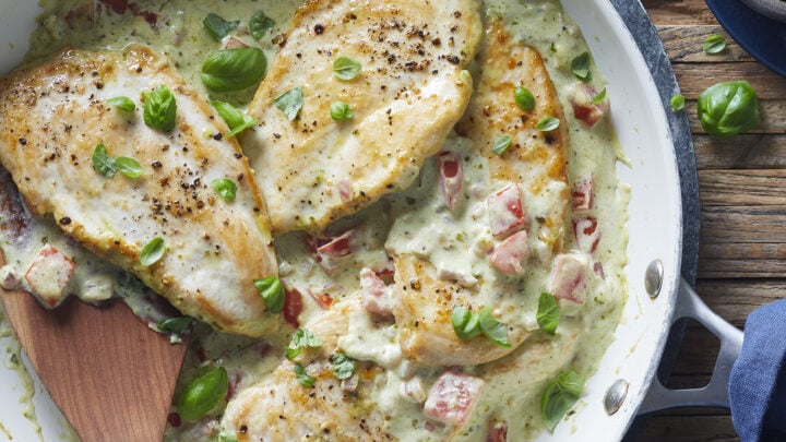 Three seared chicken breasts in a creamy sauce with diced tomatoes and fresh basil leaves in a white skillet, perfect for Healthy 20 Minute Dinners, with a wooden spatula on the side.