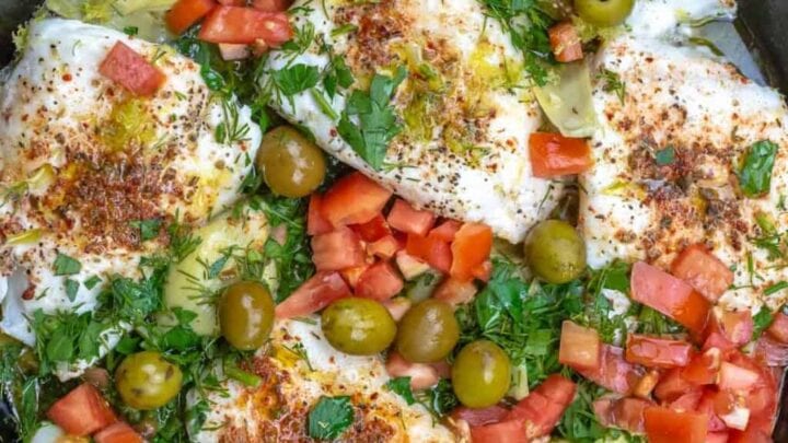 A skillet with baked white fish fillets topped with chopped tomatoes, green olives, artichoke hearts, and fresh herbs—perfect for early spring Mediterranean Diet dinners.