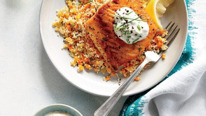 A plate with grilled fish topped with sauce and chives, served over vegetable couscous with a lemon wedge—perfect for Healthy 20 Minute Dinners; a fork, a bowl of sauce, glasses, and a napkin are nearby.