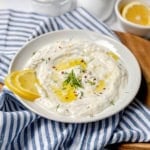 A white plate of creamy dill yogurt sauce garnished with dill, olive oil, and red pepper flakes, with two lemon slices on the side, placed on a blue-and-white striped cloth.