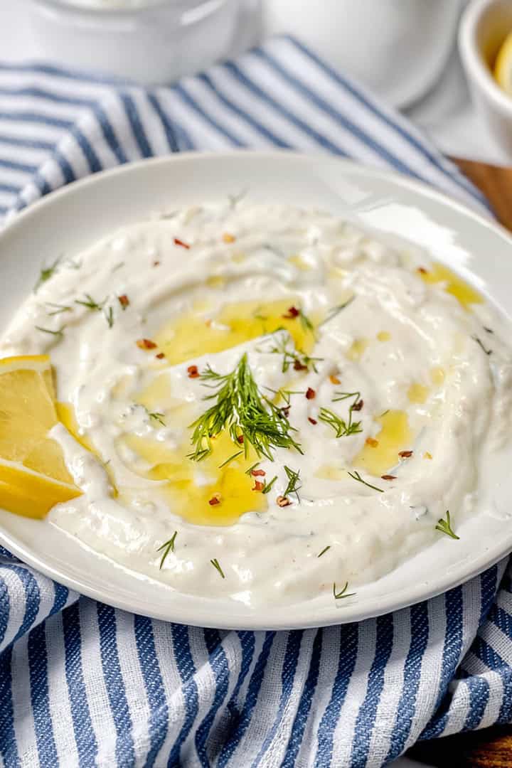A white plate of creamy dill yogurt sauce garnished with dill, olive oil, and red pepper flakes, with two lemon slices on the side, placed on a blue-and-white striped cloth.