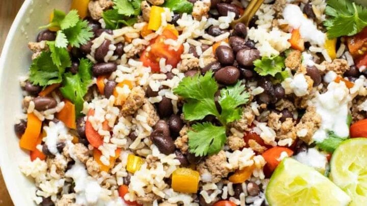 A bowl of rice topped with ground meat, black beans, chopped tomatoes, bell peppers, cilantro, lime wedges, and a drizzle of white sauce—this is one of those Healthy 20 Minute Dinners you'll love. A spoon is tucked inside for easy serving.