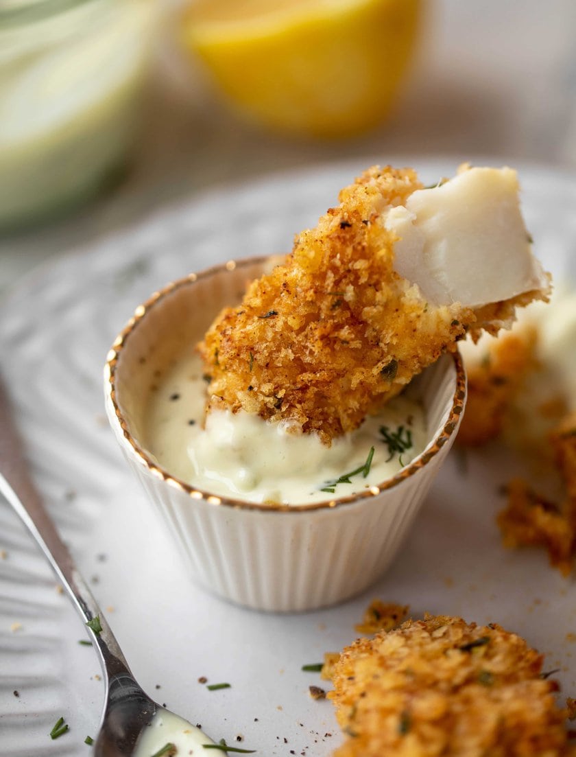 A delicious piece of breaded fish is dipped into creamy sauce, with a lemon wedge and more fish pieces in the background-perfect for those searching for tasty fish recipes for Lent.