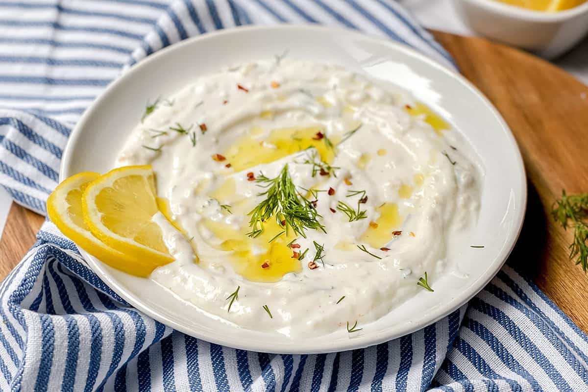 A plate of creamy Dill Yogurt Sauce garnished with olive oil, fresh dill, red pepper flakes, and lemon slices, placed on a striped cloth.