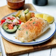 Square image of baked cod on a blue and white plate with lemon wedges and a Mediterranean salad on the side.