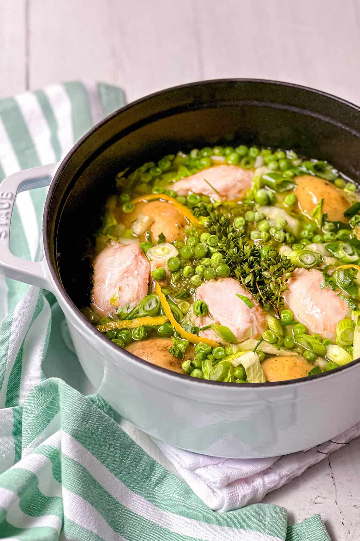 A white pot filled with Spring Chicken, potatoes, green peas, herbs, and broth sits on a striped cloth on a white surface.