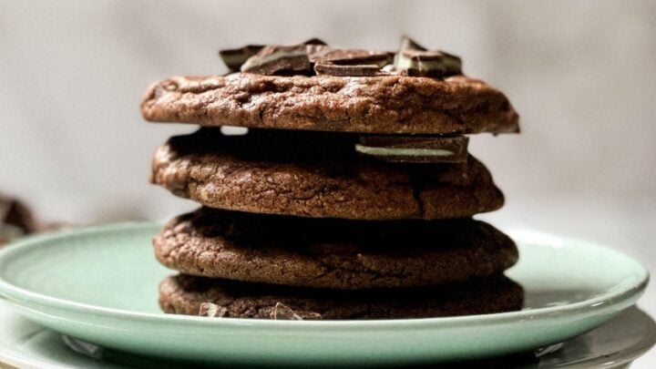 A stack of four chocolate mint cookies with chunks on top, placed on a green plate that sits on a white plate and a wooden board.