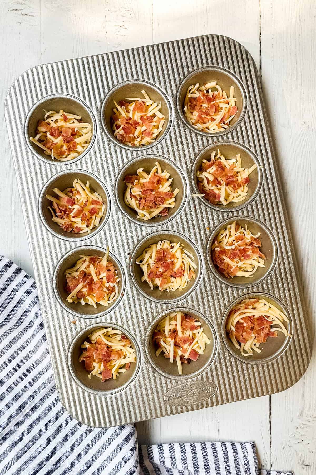 A metal muffin tin filled with shredded cheese and bits of cooked bacon in each cup, ready for your favorite egg bites recipe, sits on a white wooden surface next to a blue and white striped dish towel.