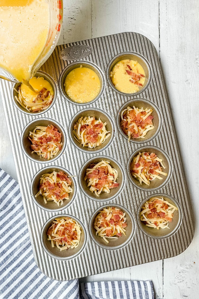 Egg custard being pour into muffin tin on top of shredded cheese and crumbled bacon.