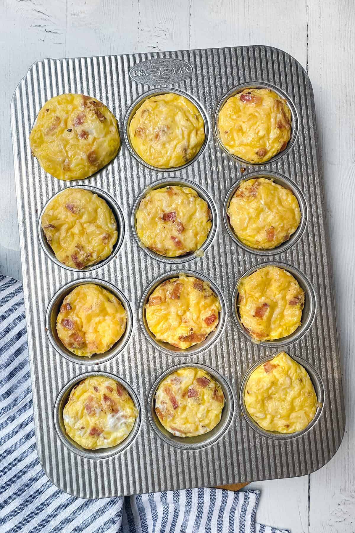 A muffin tin filled with twelve baked egg muffins, some with visible bits of ham and cheese, on a white wooden surface next to a blue and white striped cloth-perfect for trying your favorite egg bites recipe.