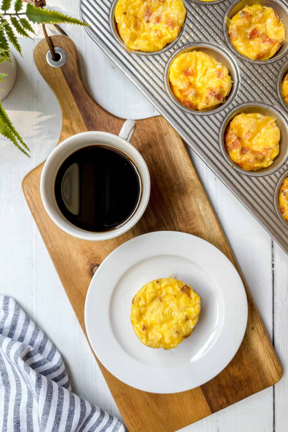 A cup of black coffee and an egg muffin on a white plate rest on a wooden board, beside a muffin tin filled with baked egg muffins-perfect for trying out your favorite egg bites recipe. A striped cloth and potted plant complete the scene.