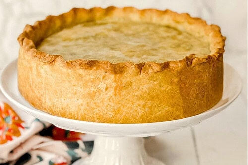 A savory Italian pizza rustica, or Easter pie, graces a white cake stand. Below, a slice reveals its rich filling of cheese and greens. A decorative floral cloth accompanies a small yellow flower nearby. Text reads "Italian Pizza Rustica Easter Pie.