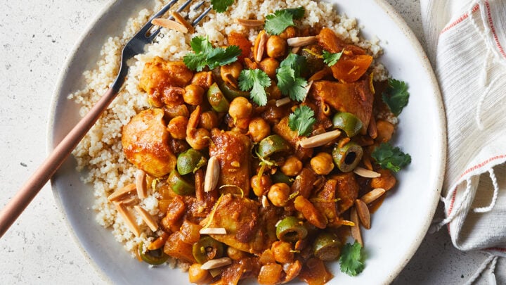 A plate of couscous topped with a chickpea and vegetable stew, garnished with cilantro and slivered almonds—a perfect March 400 Calorie Dinners option—served with a fork and a glass of water.
