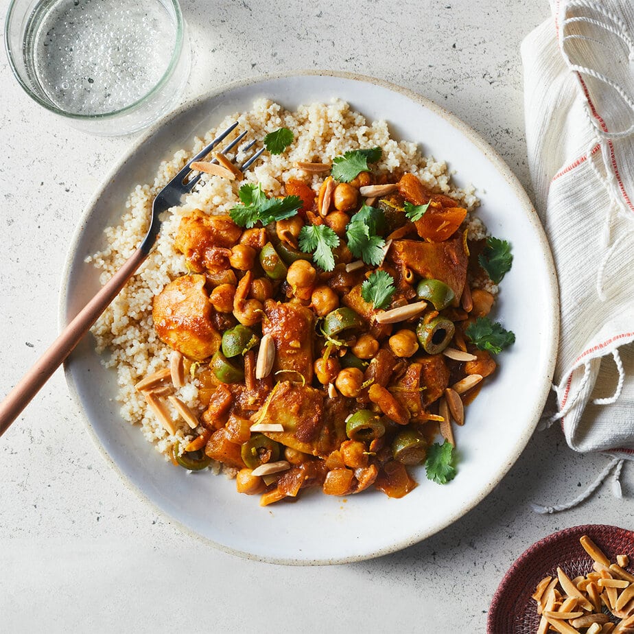 A plate of couscous topped with a chickpea and vegetable stew, garnished with cilantro and slivered almonds-a perfect March 400 Calorie Dinners option-served with a fork and a glass of water.