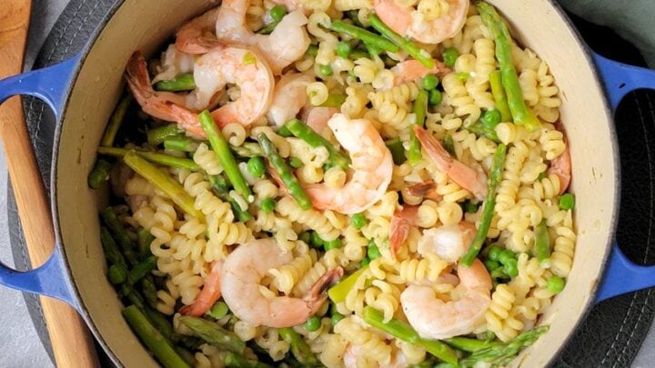 A pot of spiral pasta mixed with shrimp and asparagus sits on a gray surface next to a wooden spoon and a green cloth—perfect for easy March One Pot Dinners.