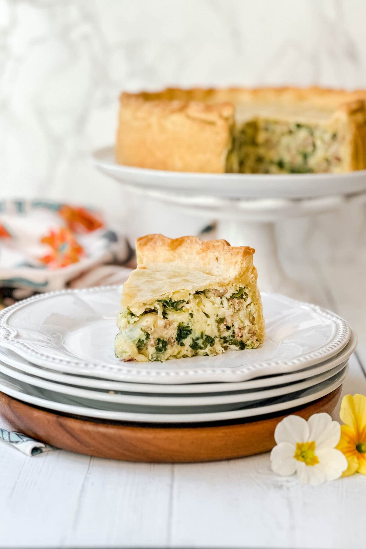 A slice of pizza rustica on a stack of white plates, showcasing a golden crust filled with spinach and a creamy mixture. The rest of the pie rests elegantly on a stand in the background, while floral decor peeks out beside the plates.