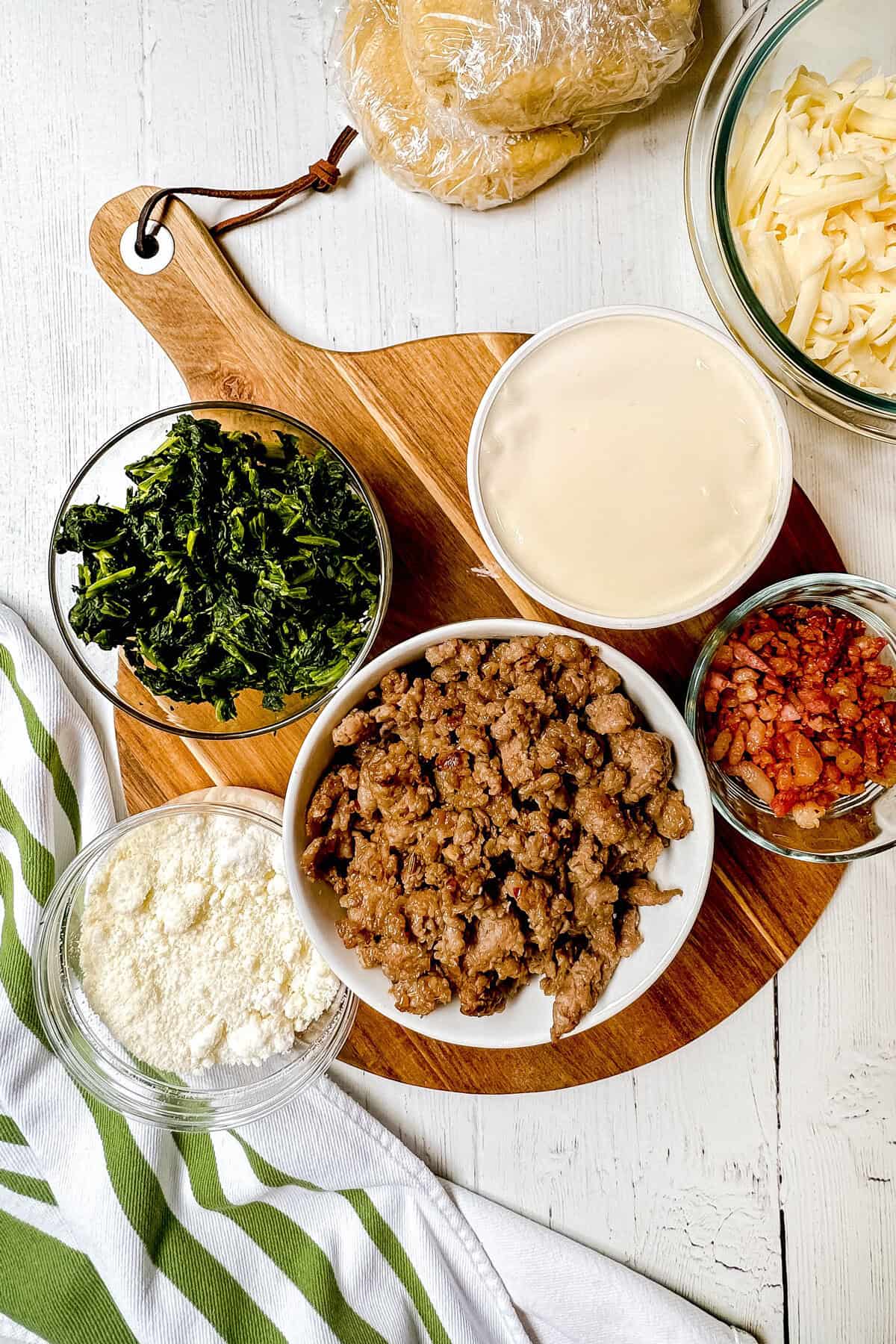 A wooden board displays bowls of cooked sausage, chopped kale, white sauce, bacon bits, grated cheese, and crumbled cheese on a white table with a striped towel. In the background, dough wrapped in plastic hints at delightful pizza rustica preparations.