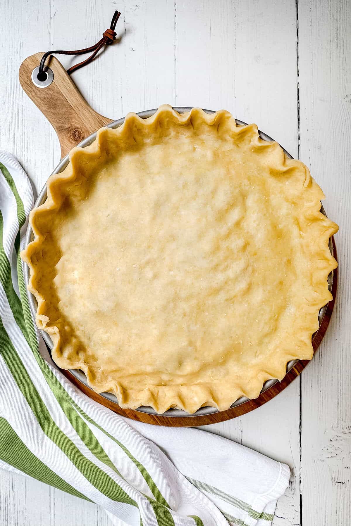 A pie crust in a fluted dish rests on a wooden board, set on a white wooden surface. A green and white striped cloth lies nearby, evoking the rustic charm of pizza rustica.